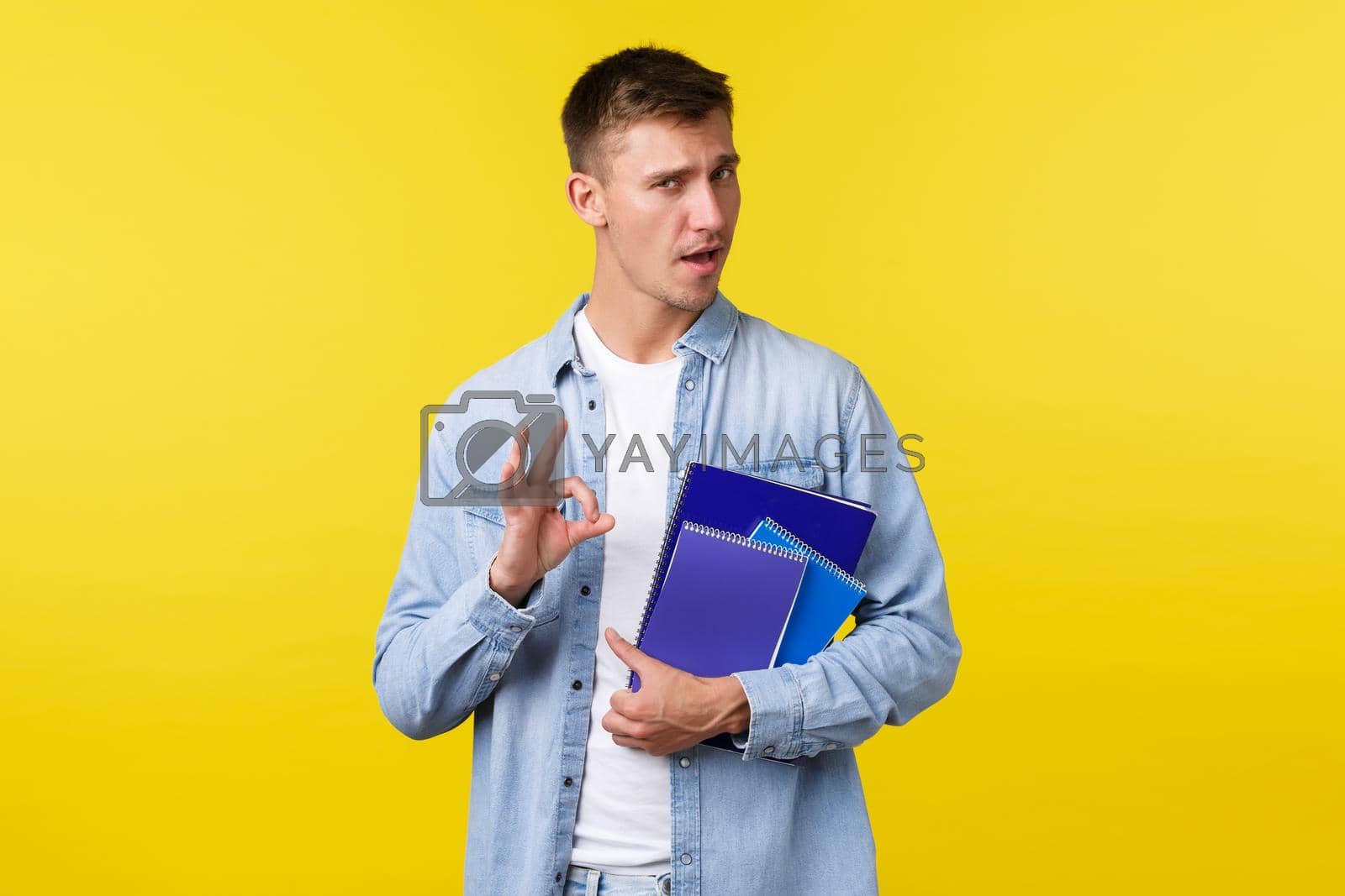 Royalty free image of Education, courses and university concept. Handsome assured blond guy encourage enroll this college, showing okay gesture as guarantee you will like it, holding notebooks, yellow background by Benzoix