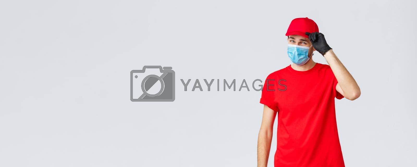 Royalty free image of Covid-19, self-quarantine, online shopping and shipping concept. Sassy delivery man in red uniform and mask with gloves, saluting client, deliver packages and orders during coronavirus, food orders by Benzoix
