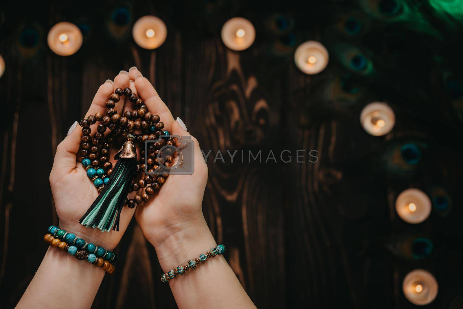 Royalty free image of Woman holds in hand wooden mala beads strands used for keeping count during mantra meditations. Weaving and creation. Wooden background with candles and feathers. Spirituality, religion, God concept. by kristina_kokhanova