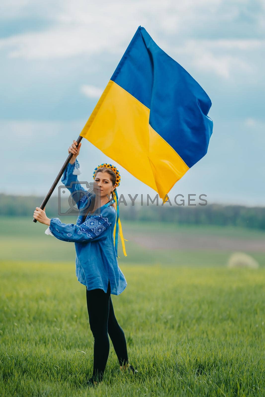Royalty free image of Beautiful ukrainian woman with national flag on green field background. Young lady in blue embroidery vyshyvanka. Ukraine, independence, freedom, patriot symbol, victory in war. by kristina_kokhanova
