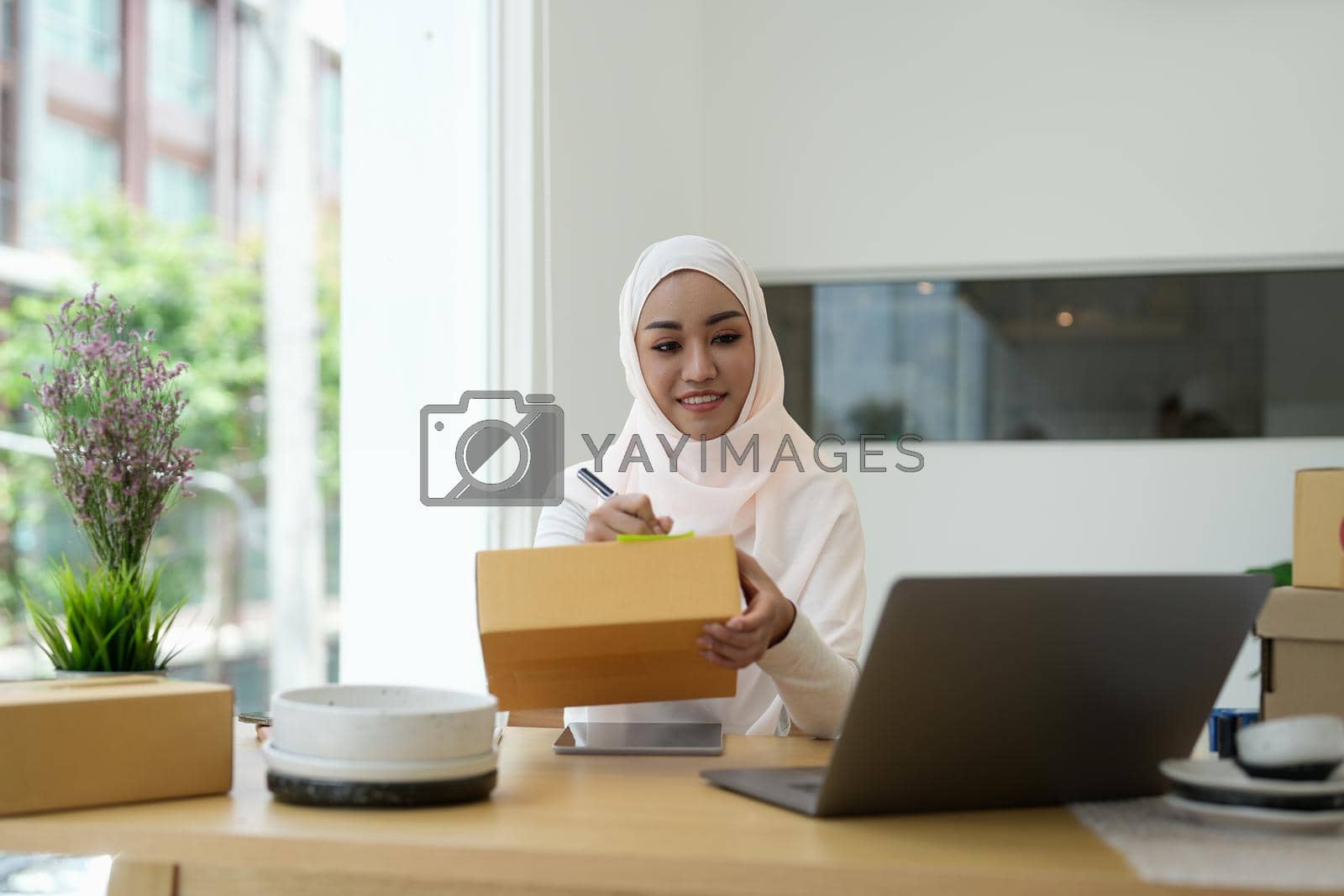 Royalty free image of Muslim female online store small business owner seller entrepreneur packing package post shipping box preparing delivery parcel on table. Ecommerce dropshipping shipment service concept. by nateemee