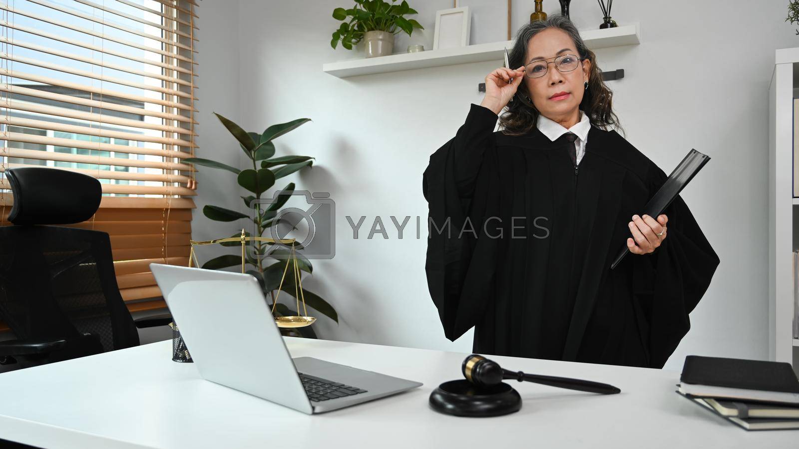 Royalty free image of Positive mature female judge, lawyer or attorney in robe gown uniform standing at her personal office. Law and justice concept by prathanchorruangsak