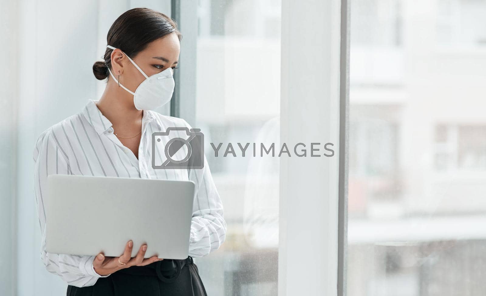 Royalty free image of A chance to do things differently. a masked young businesswoman holding a laptop and looking out a window in a modern office. by YuriArcurs