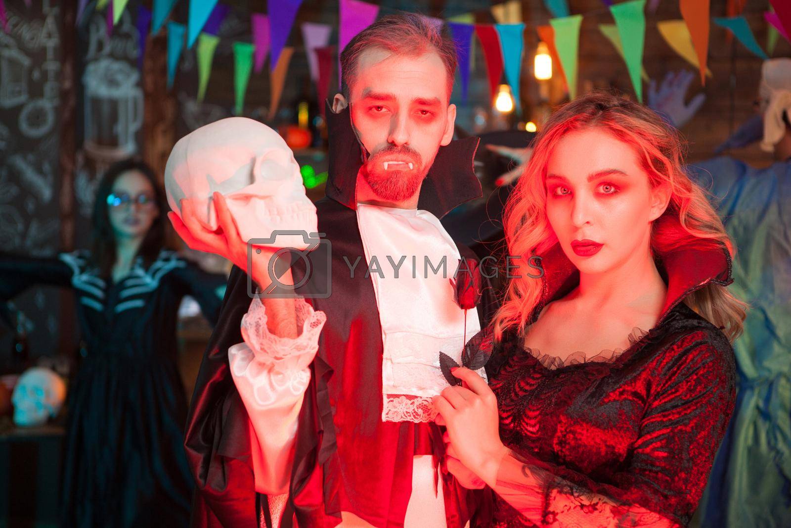 Portrait of terrifying zombie couple at halloween celebration. Man dressed up like dracula holding a human skull.