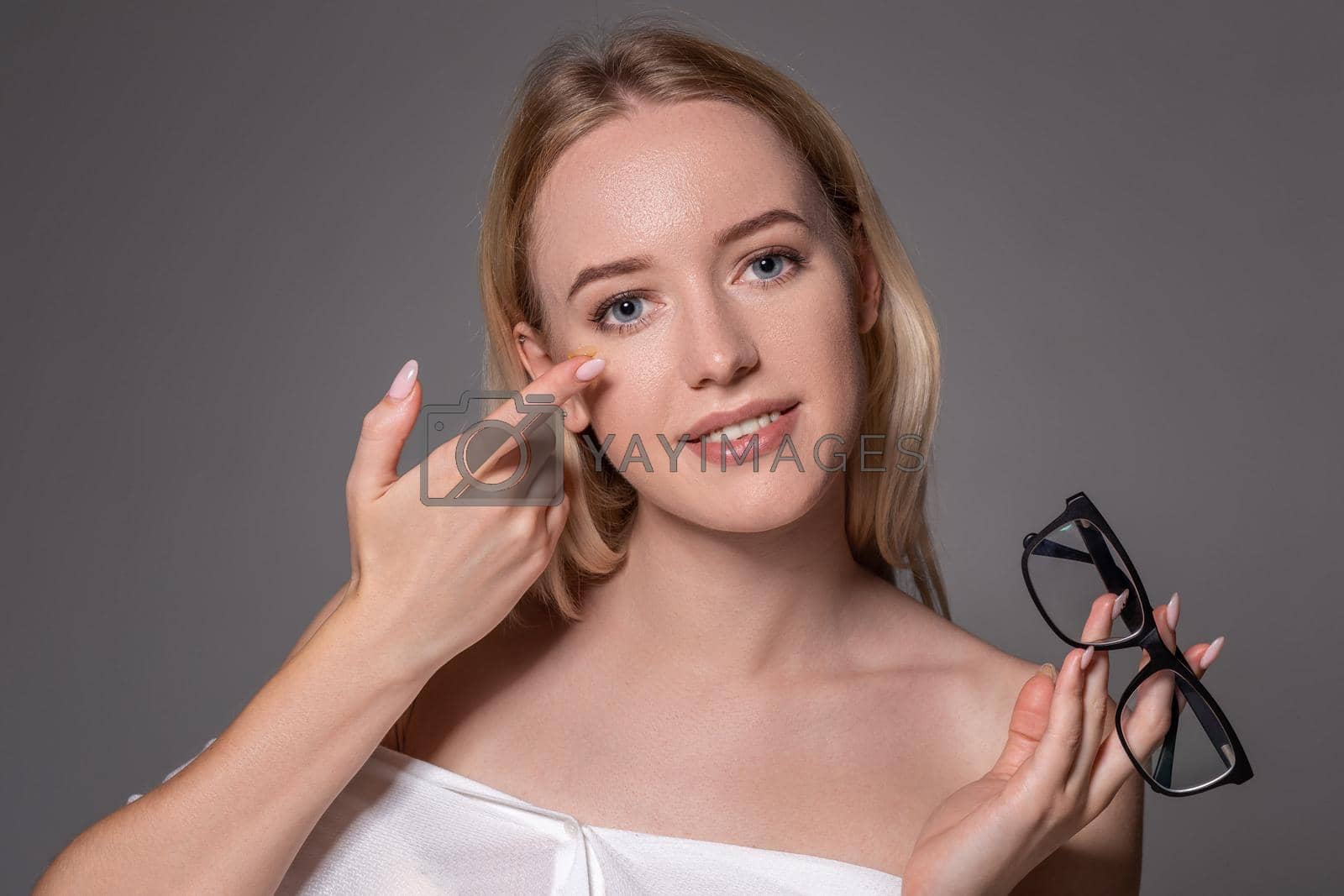 Royalty free image of Young blonde woman holding contact lens on finger in front of her face and holding in her other hand a black glasses on gray background. by nazarovsergey