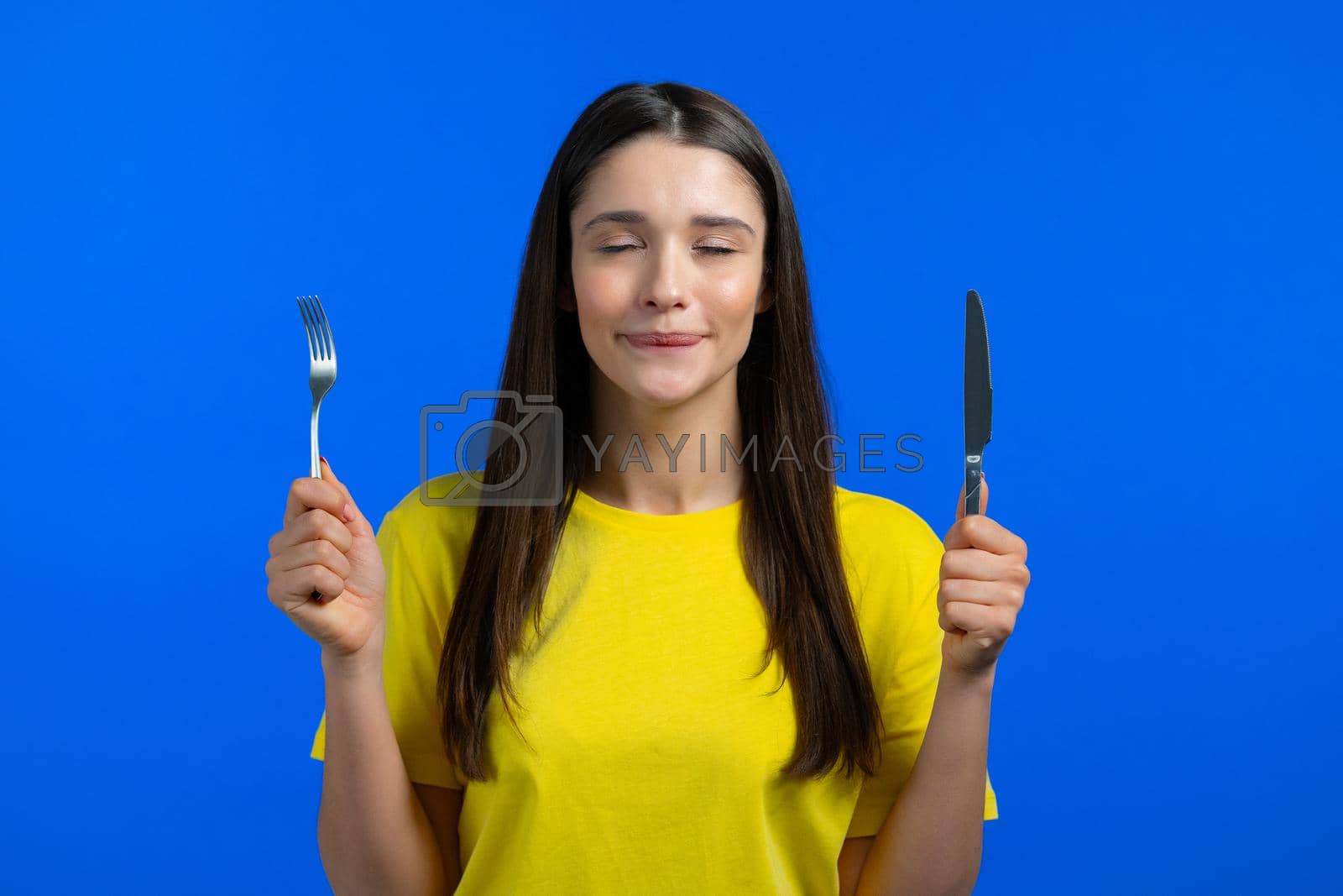 Royalty free image of Portrait of hungry woman with fork and knife. Lady with anticipation waiting for serving dinner dishes with cutlery on blue studio background. by kristina_kokhanova