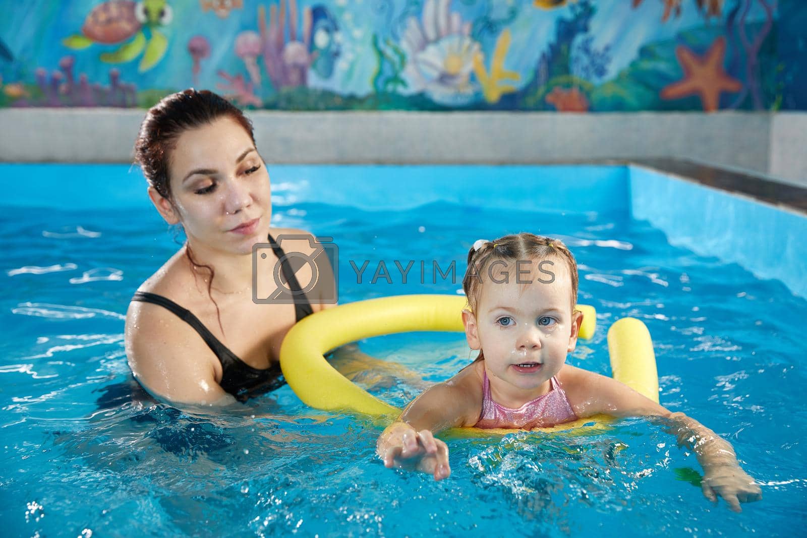 Royalty free image of Preschool girllearning to swim in pool with foam noodle with young trainer by Mariakray