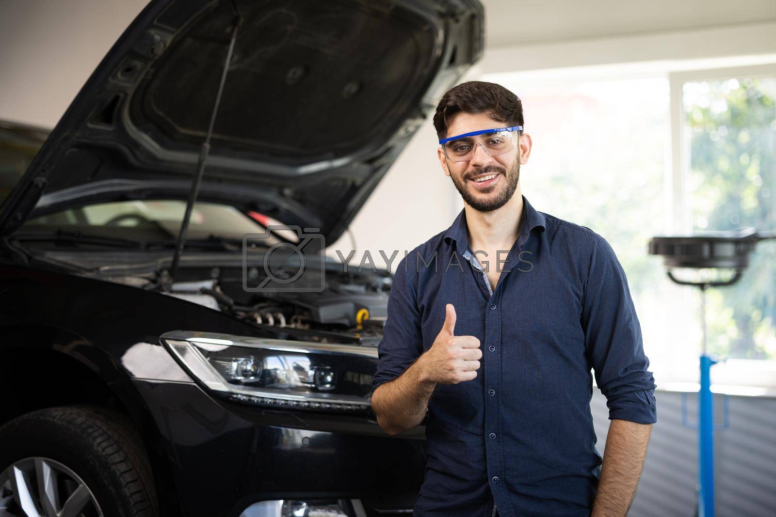 Royalty free image of Happy technician bearded brunet mechanic man 20s in safety glasses work in vehicle repair shop show thumb up like gesture on light workshop background indoors by uflypro