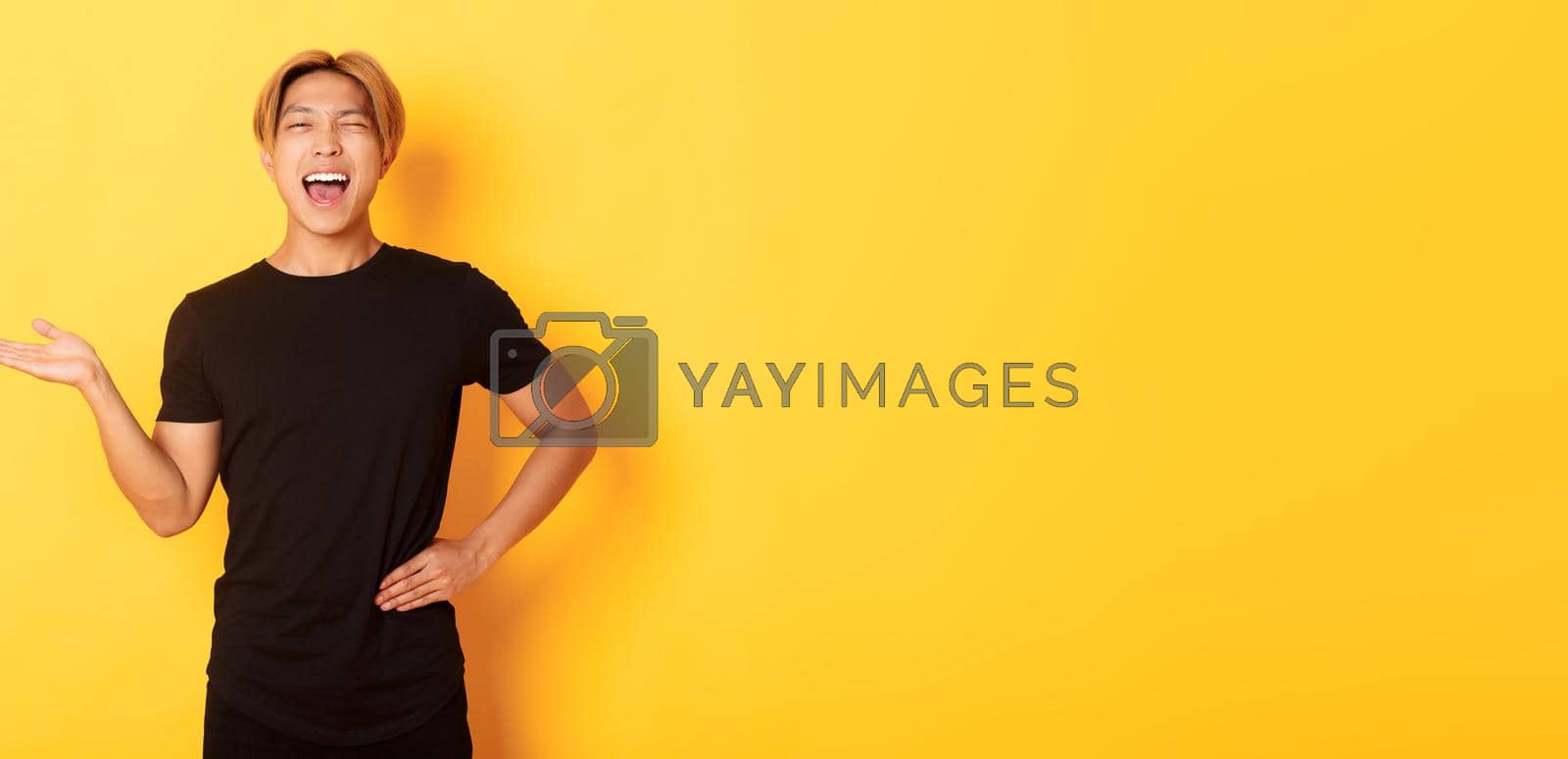 Royalty free image of Portrait of happy blond asian guy, winking sassy and smiling, holding something on hand over yellow background by Benzoix