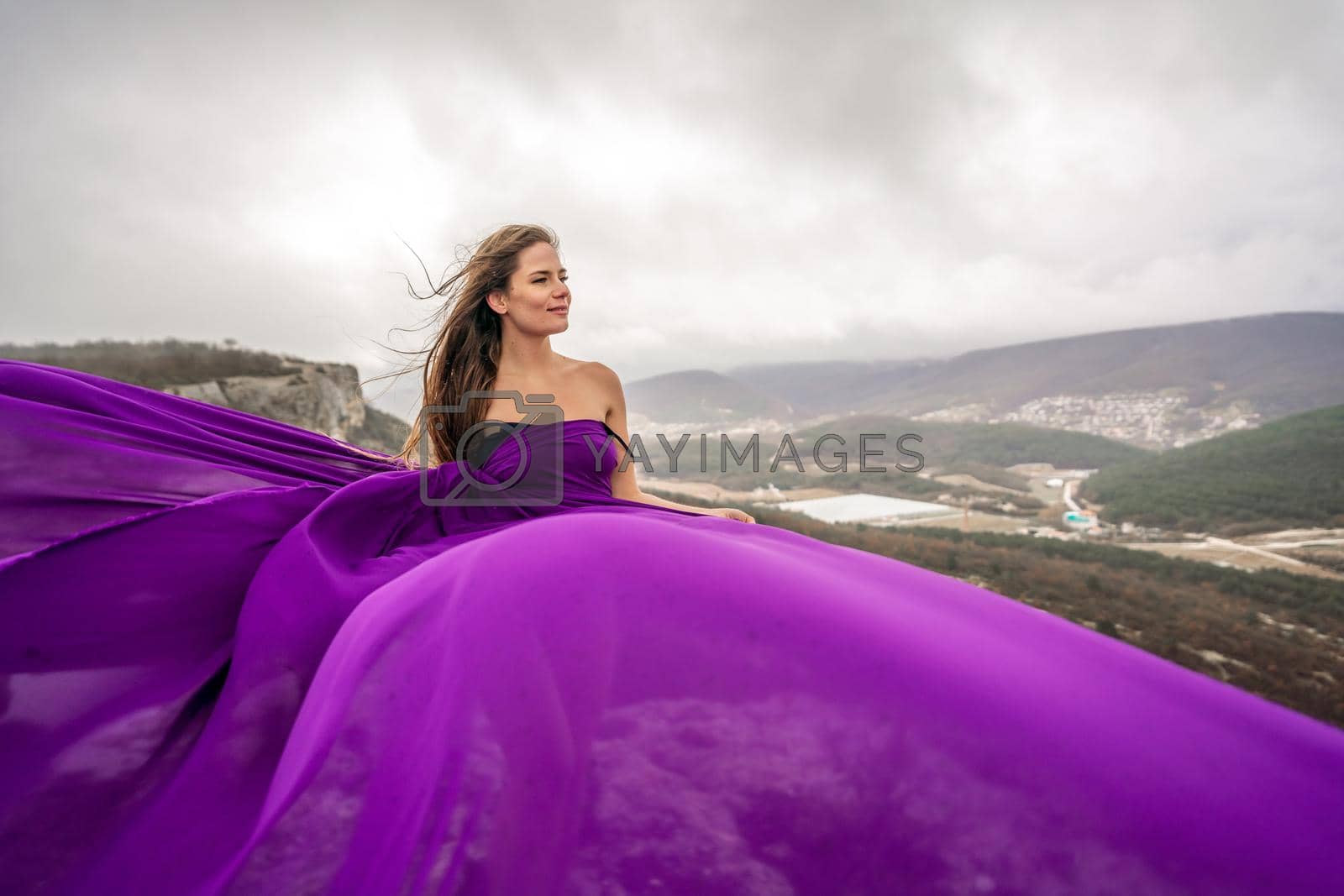 Royalty free image of A woman with long hair is standing in a purple flowing dress with a flowing fabric. On the mountain against the background of the sky with clouds. by Matiunina