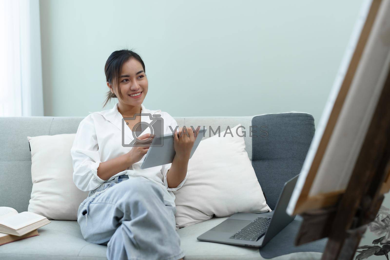 Royalty free image of Portrait of an Asian woman using a tablet to paint artwork on a sofa. by Manastrong
