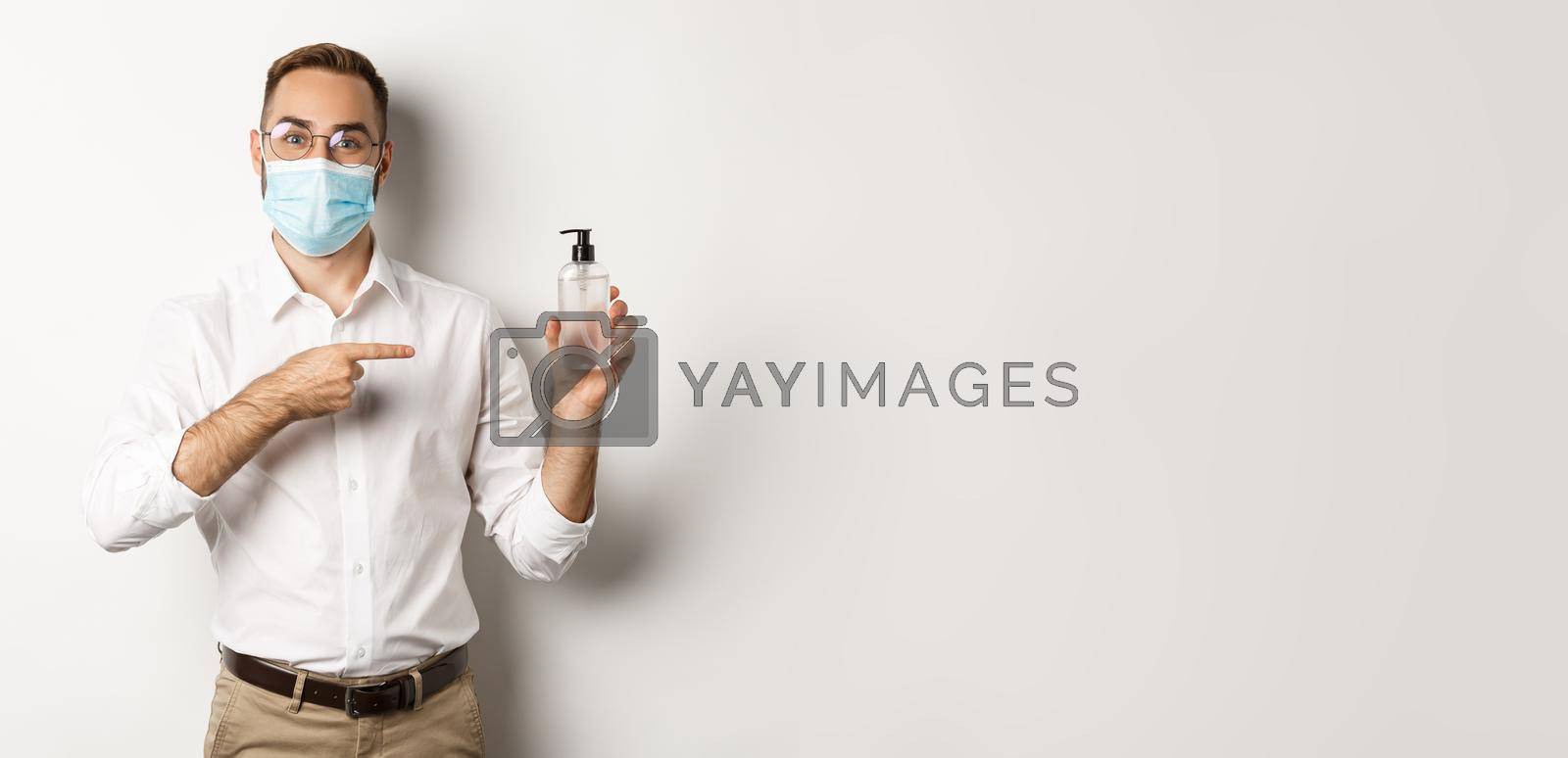 Royalty free image of Covid-19, social distancing and quarantine concept. Office worker in medical mask pointing at hand sanitizer, showing antiseptic, white background by Benzoix