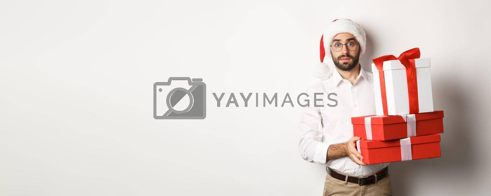 Royalty free image of Merry christmas, holidays concept. Confused man in Santa hat holding pile of presents, found gifts under xmas tree, standing against white background by Benzoix