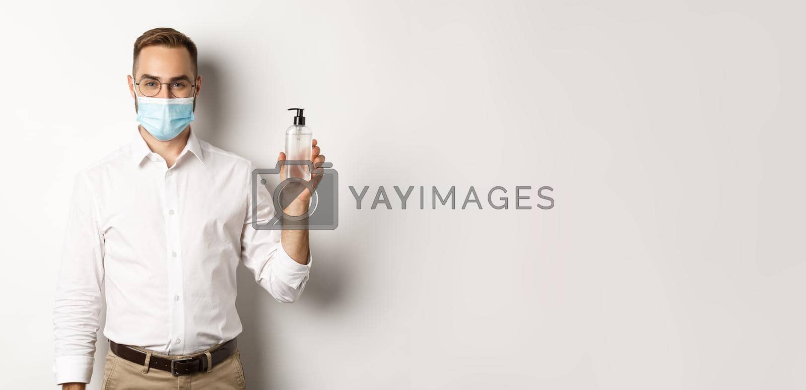 Royalty free image of Covid-19, social distancing and quarantine concept. Employer in medical mask showing hand sanitizer, asking to use antiseptic at work, standing over white background by Benzoix