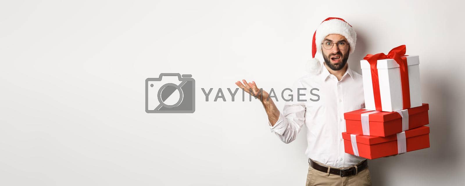 Royalty free image of Merry christmas, holidays concept. Man looking confused while holding xmas gifts, shrugging puzzled, standing in santa hat against white background by Benzoix