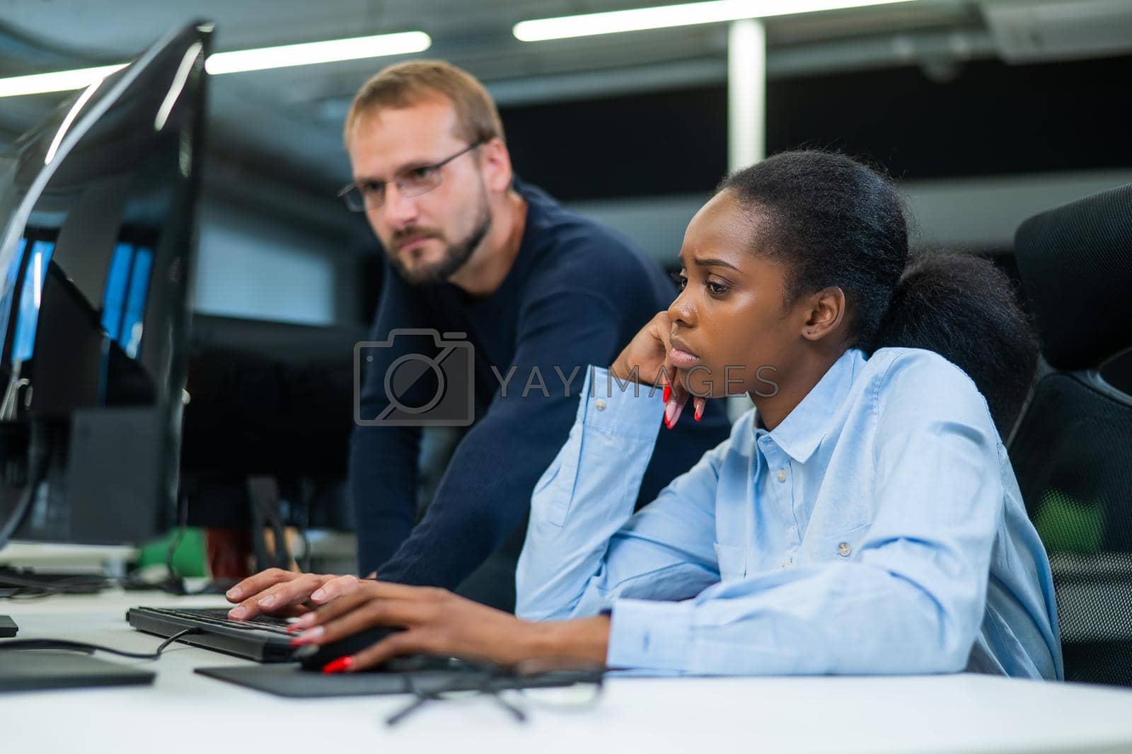 Royalty free image of Colleagues look at the monitor and decide working moments. Caucasian man helps sad african woman solve computer problem. by mrwed54