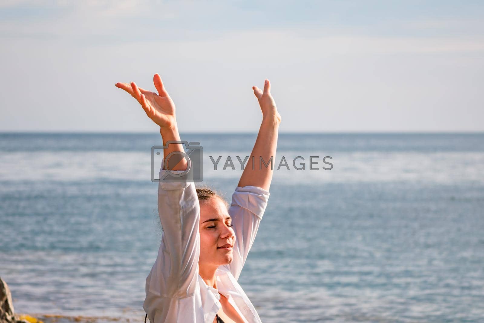 Royalty free image of The girl stands on the shore and looks at the sea. Her hands are raised up. She wears a white shirt and her hair is in a braid. by Matiunina