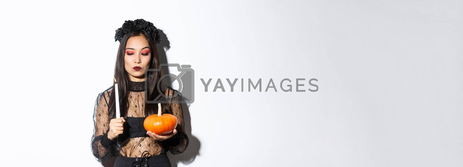 Royalty free image of Myserious asian wicked witch in gothic dress, looking at lit candle, holding pumpkin, standing over white background by Benzoix