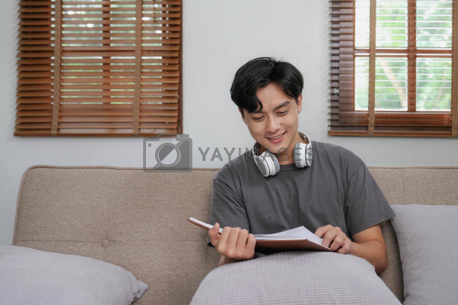 Royalty free image of Handsome Asian man relaxing on the sofa at home with a book and headphones by wichayada