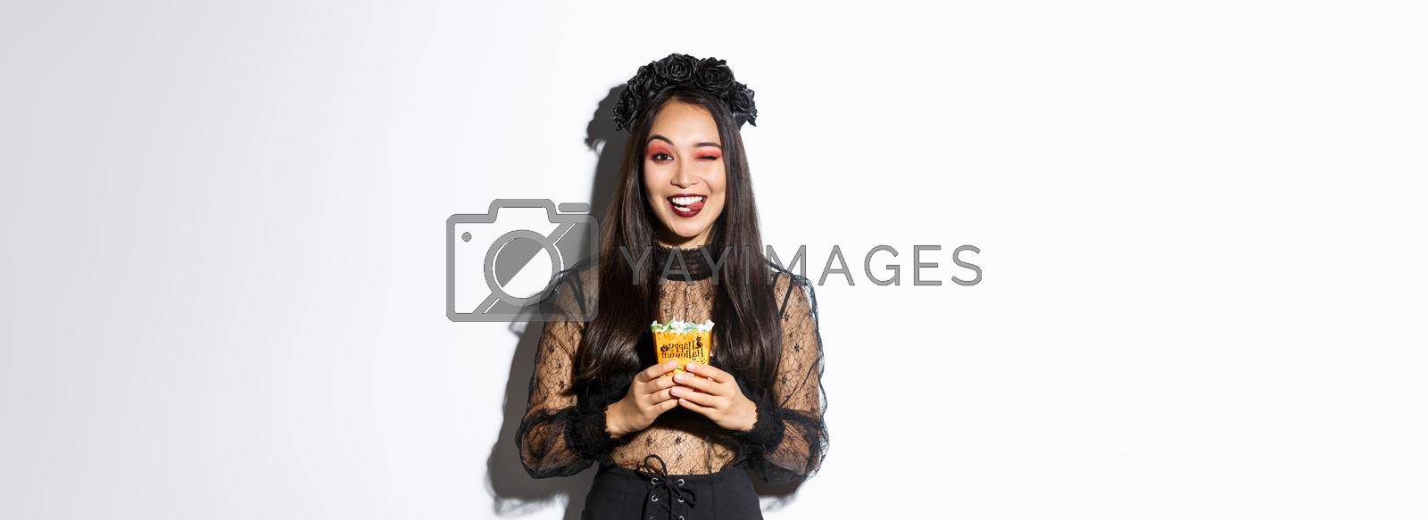 Royalty free image of Cheeky smiling girl in witch costume, celebrating halloween, going trick or treat in gothic dress, showing tongue and holding sweets by Benzoix