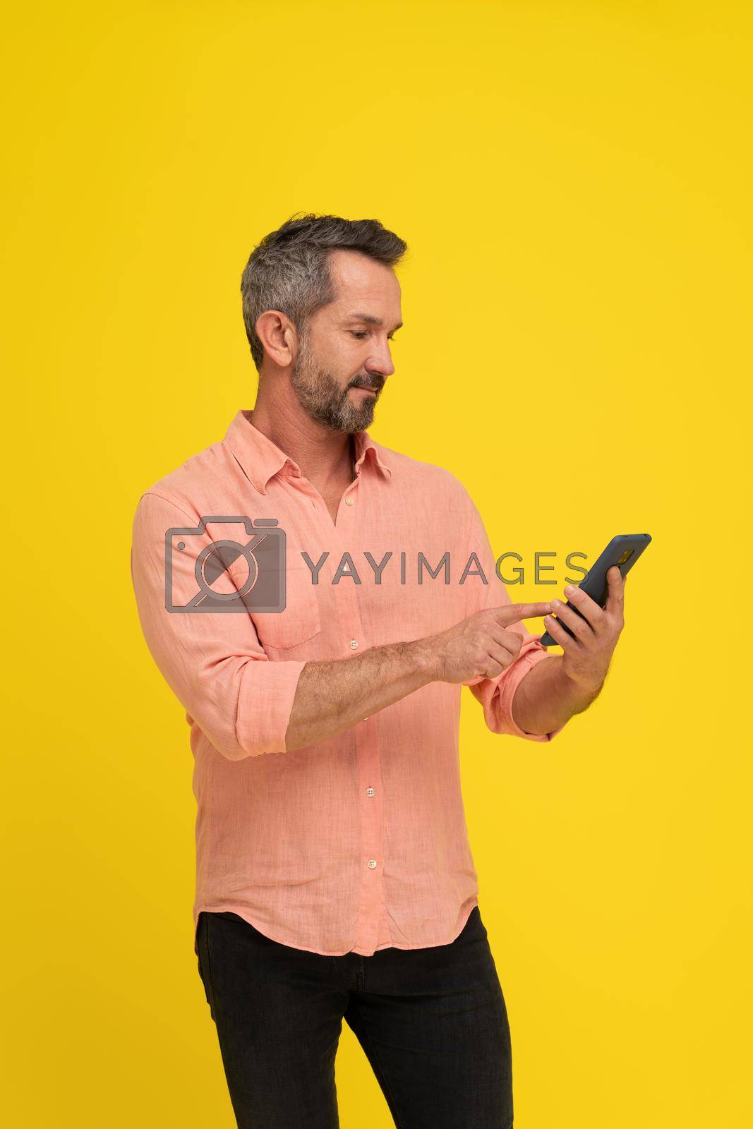 Royalty free image of Masculine, fit middle aged grey haired handsome man holding smartphone looking at it wearing peach shirt isolated on yellow. Mature muscled man with phone, mobile app advertisement by LipikStockMedia