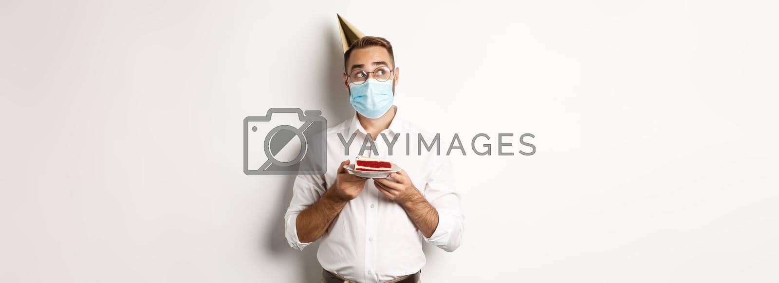 Royalty free image of Covid-19, social distancing and celebration. Thoughtful man holding birthday cake, making wish and wearing face mask on quarantine, white background by Benzoix