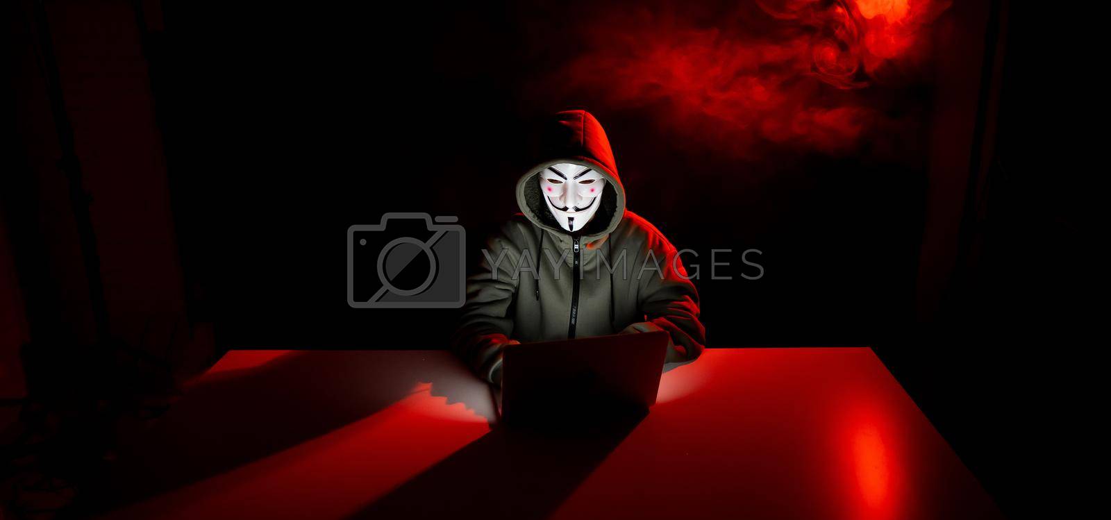 Royalty free image of Anonymous in a hood is typing on a laptop in the dark in red smoke. by mrwed54