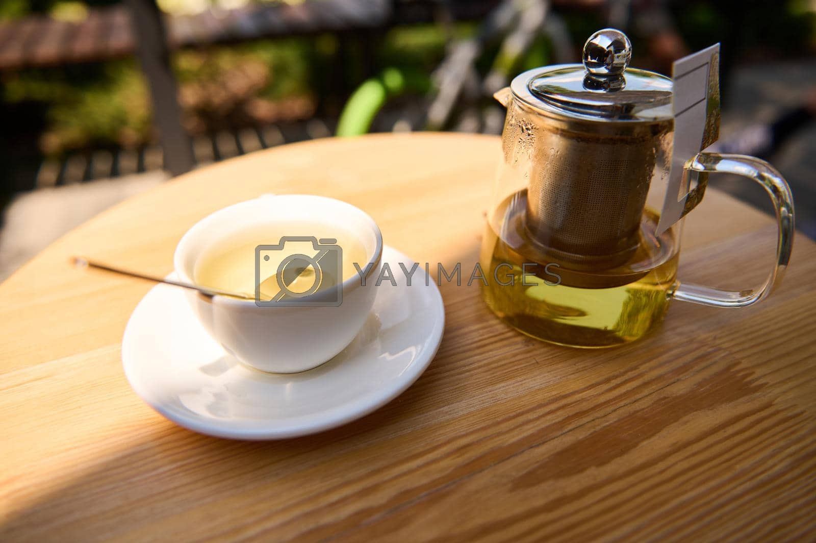 Close-up of a glass teapot and white cup with healthy green herbal tea, on a saucer, on a wooden table in the summer terrace. Food and drink consumerism