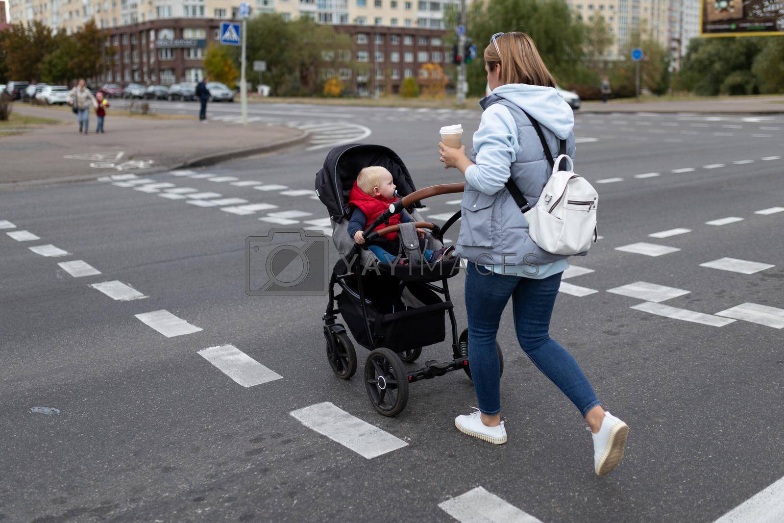 Royalty free image of a young mother with a stroller crosses the road at a pedestrian crossing with a cup of coffee in her hands by TRMK