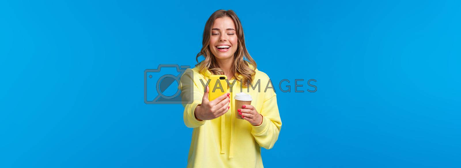 Royalty free image of Cheerful smiling blond caucasian female blogger record video or take selfie on her smartphone, laughing and grinning as holding take-away cup of coffee, stand blue background by Benzoix