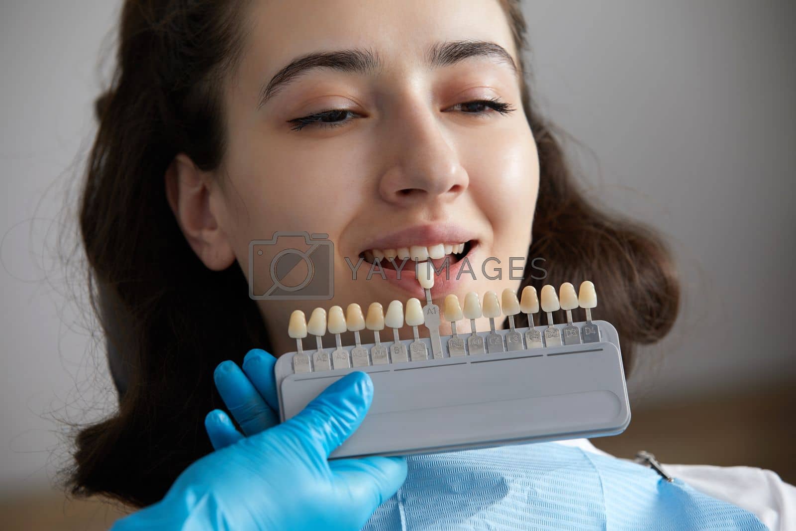 Royalty free image of Dentist choosing color of tooth enamel for patient. Dentist applying sample from tooth enamel scale to caucasian female teeth by Mariakray
