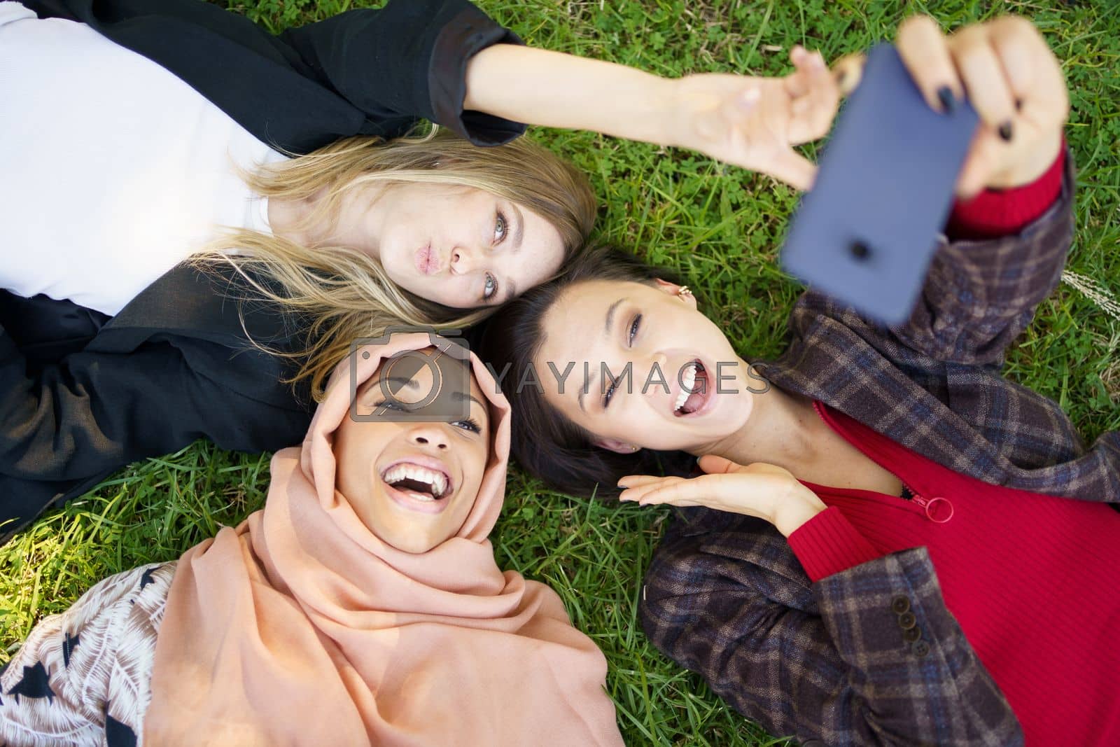Royalty free image of Multiracial girlfriends laughing while lying on lawn and taking selfie by javiindy