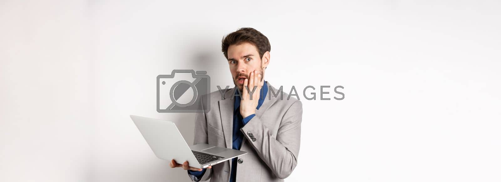 Royalty free image of Shocked and troubled businessman have trouble on laptop, looking worried at camera, made mistake at work, standing on white background in suit by Benzoix