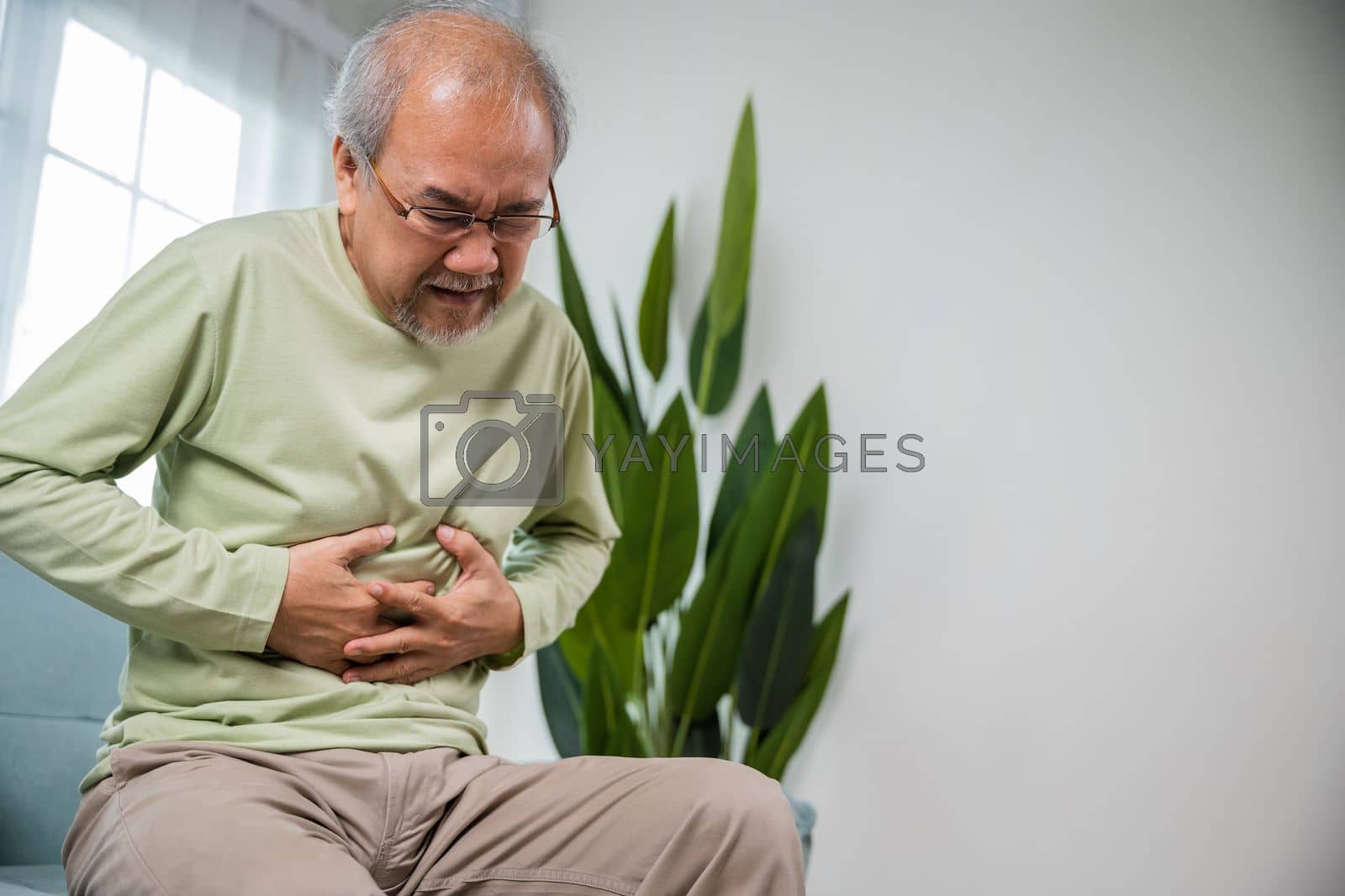 Royalty free image of Senior Asian man sitting on sofa having suffering from stomach ache holding his stomach pain by Sorapop