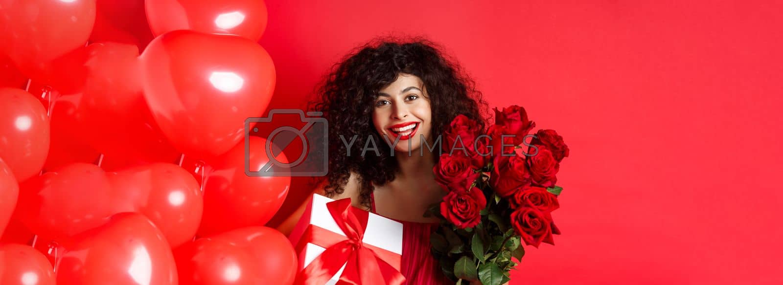 Royalty free image of Happy Valentines day. Romantic girl with presents from lover, holding bouquet of roses and gift box, standing near cute red hearts on studio background by Benzoix