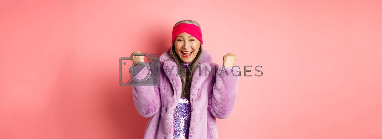 Royalty free image of Cheerful asian senior woman winning prize and celebrating. Old funky lady saying yes and clench fists in triumph, standing over pink background by Benzoix