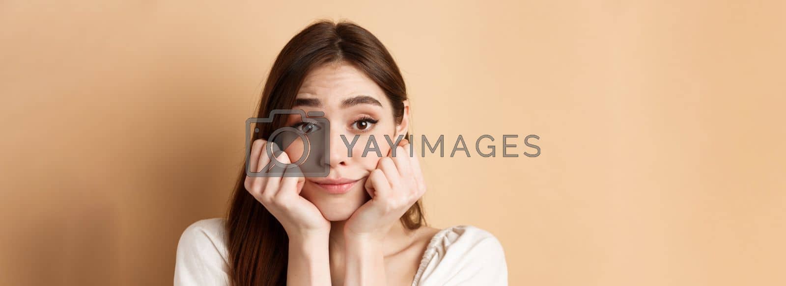 Royalty free image of Close-up of cute and pretty girl lean face on hands, looking at camera with dreamy smile, standing on beige background by Benzoix