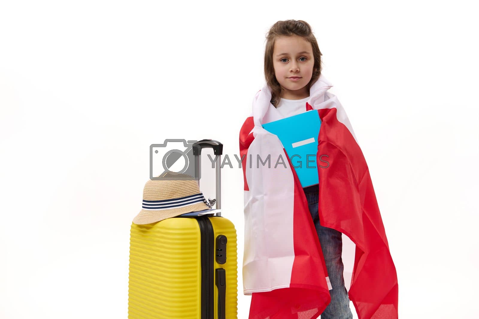 Royalty free image of Happy first grader, little girl wrapping in Canada flag, holding a copybook, posing with suitcase over white background. by artgf