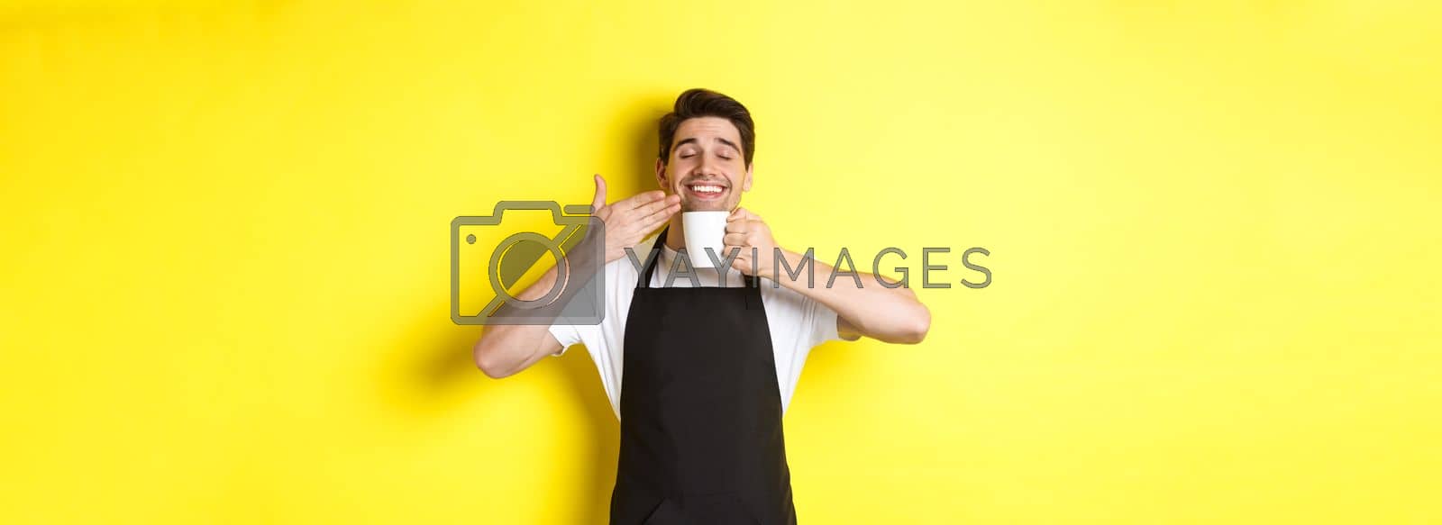 Royalty free image of Barista enjoying smell of coffee in mug, standing pleased with eyes closed, wearing black apron by Benzoix