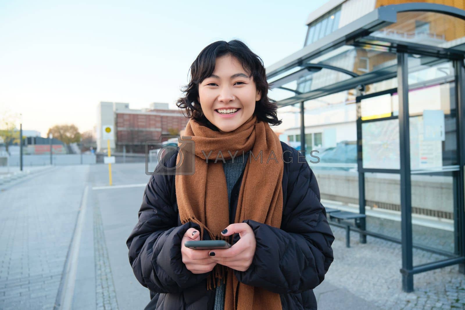 Royalty free image of Portrait of Korean woman in winter jacket, standing with smartphone, waiting for bus on stop, looking at mobile phone app checking public transport application by Benzoix