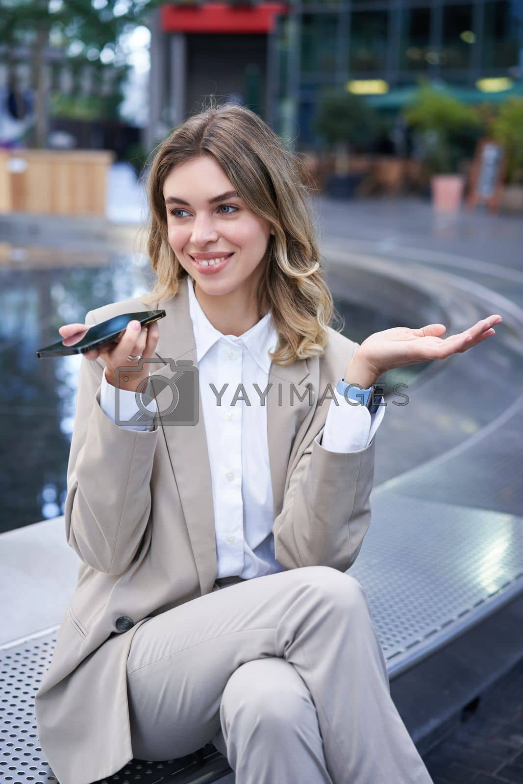 Royalty free image of Smiling business woman record voice message, speaking into microphone on mobile phone, sitting near fountain on street by Benzoix