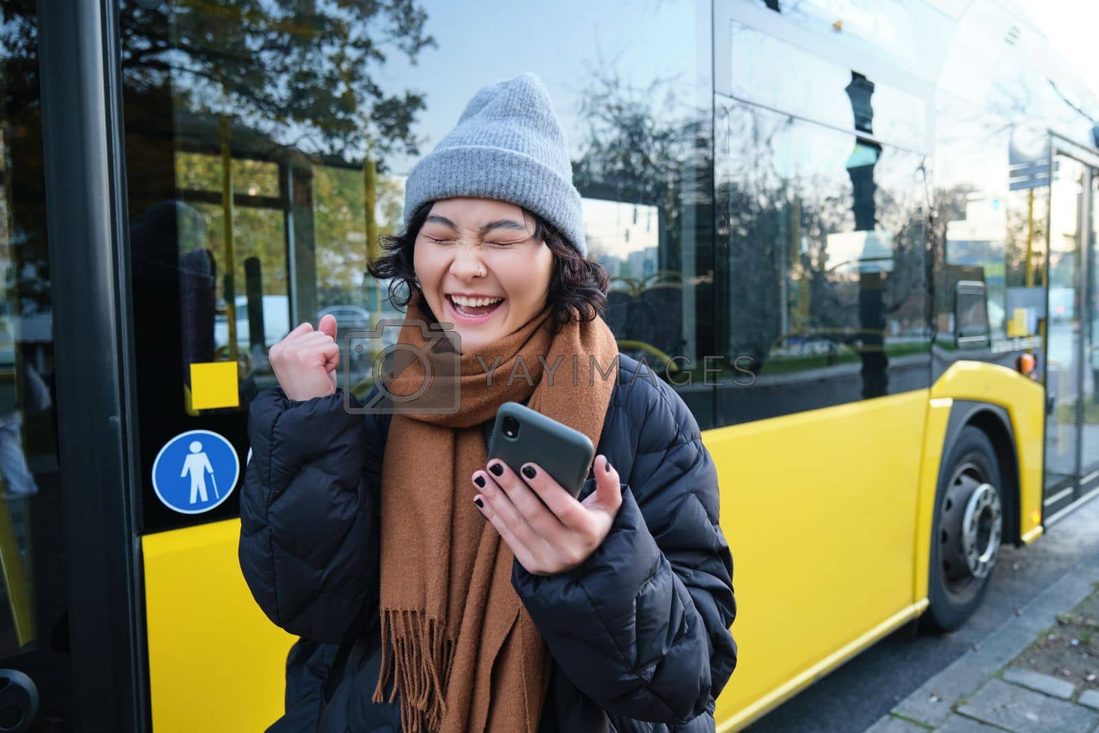 Royalty free image of Modern people and lifestyle. Happy asian girl screams from joy, celebrates, stands near bus public transport and looks amazed by Benzoix