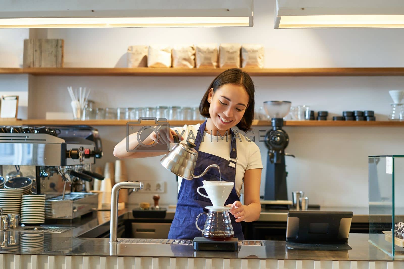 Royalty free image of Smiling asian girl barista, pouring hot water from kettle, brewing filter coffee, standing behind counter in cafe by Benzoix