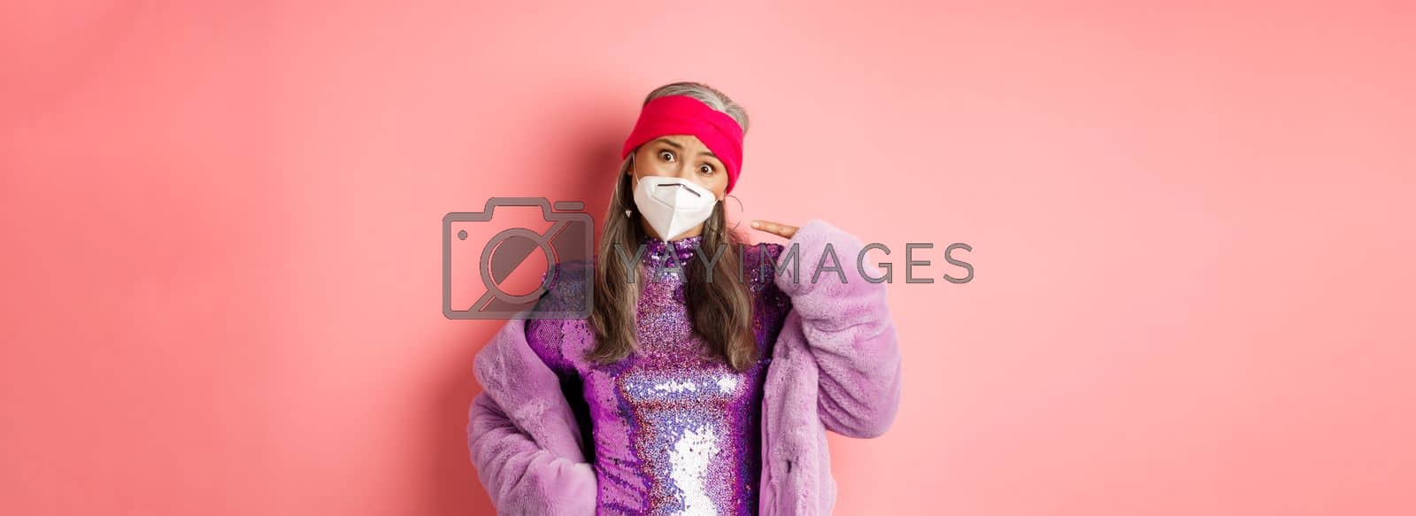 Royalty free image of Covid-19, pandemic and fashion concept. Asia senior woman in stylish glittering dress complaining on face mask, pointing at respirator and frowning, pink background by Benzoix