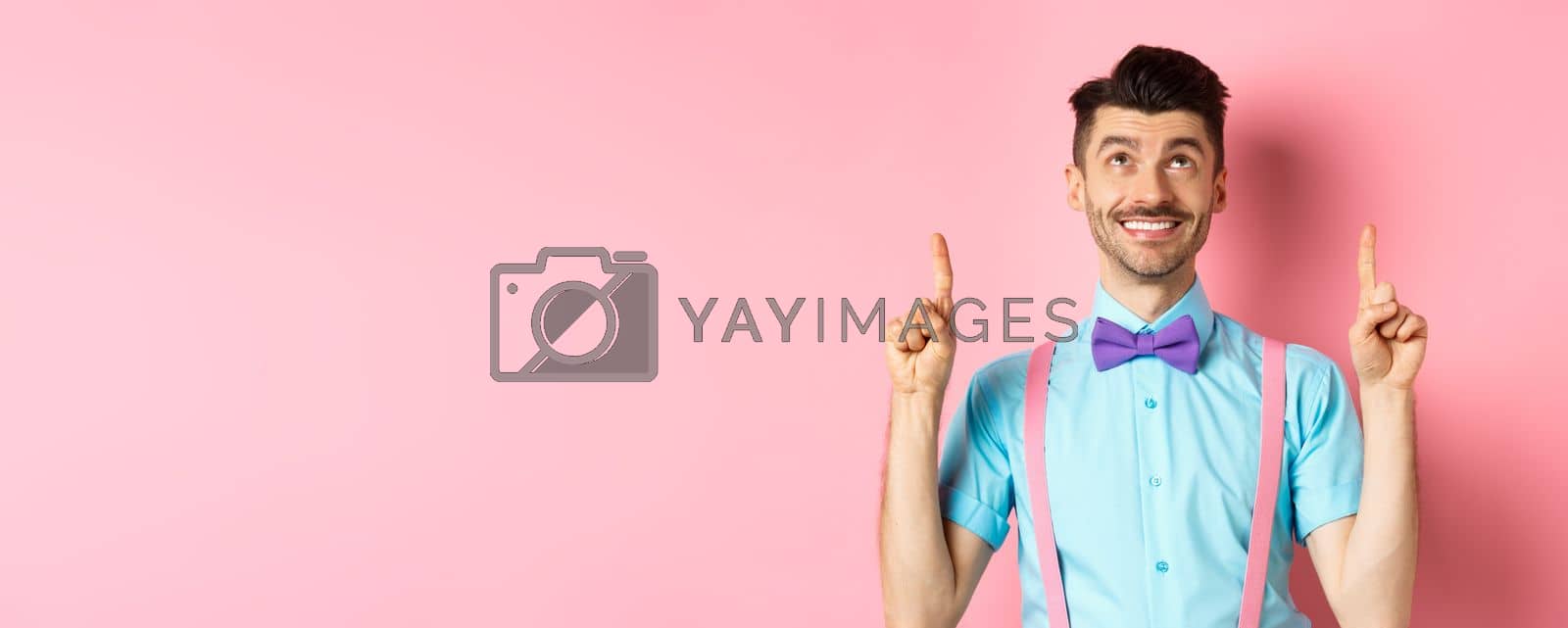 Royalty free image of Happy smiling man in bow-tie pointing, looking up, showing something cool, standing on pink background by Benzoix