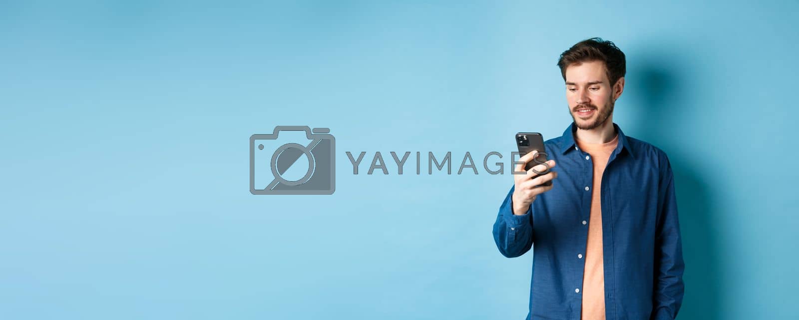 Royalty free image of Handsome modern guy using mobile phone, reading smartphone screen and smiling, networking on blue background by Benzoix
