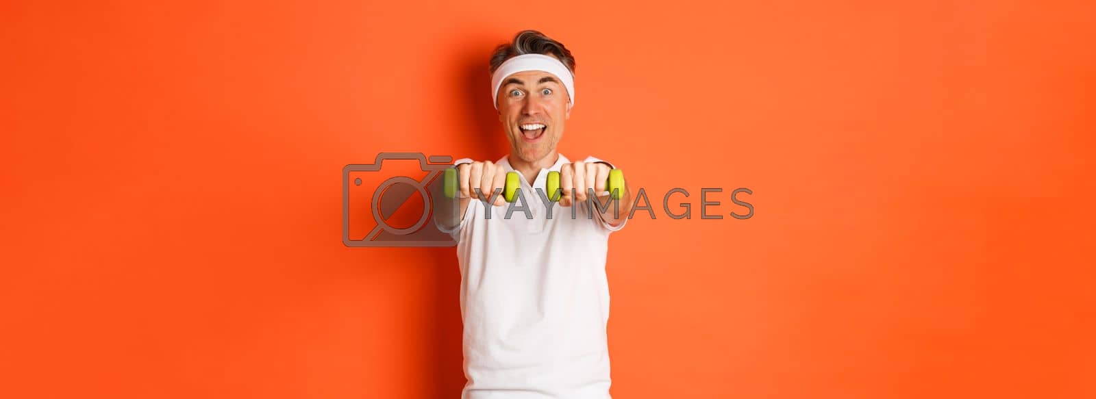 Image of active middle-aged fitness guy, doing sport exercises with dumbbells and smiling excited, standing over orange background.