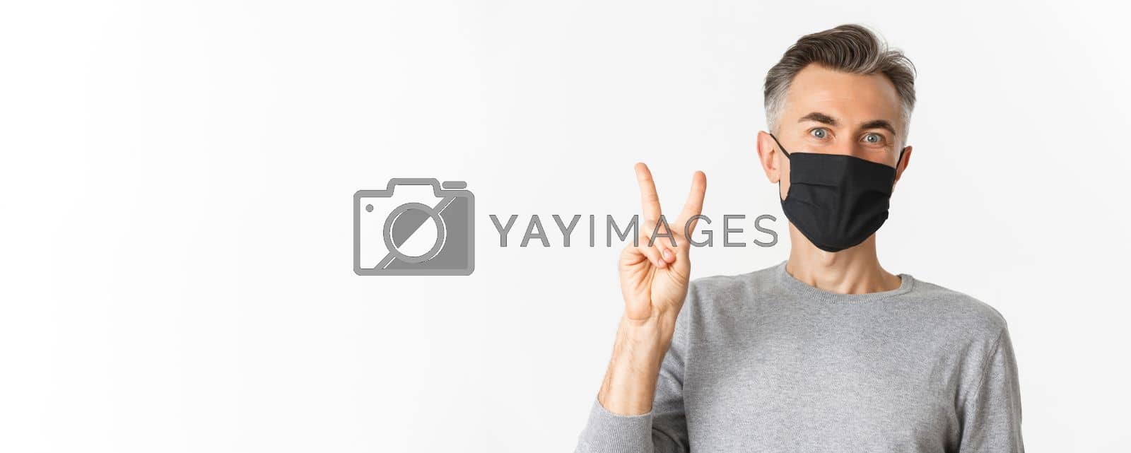 Royalty free image of Concept of covid-19, social distancing and quarantine. Close-up of cheerful middle-aged guy in medical mask, showing peace sign and smiling, standing over white background by Benzoix