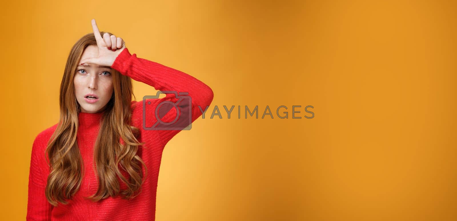 Royalty free image of Arrogant and self-satisfied redhead woman humiliating person showing loser sign on forehead mocking and disdain rival standing confident and snobbish over orange background by Benzoix