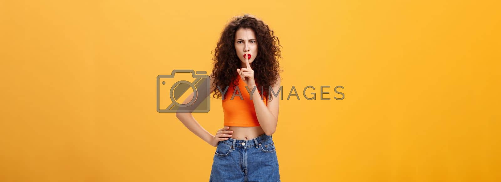 Royalty free image of Sister displeased sibling saying cursing words standing serious and strict over orange background in stylish outfit shushing at camera saying shh with index finger over mouth demanding obey rules by Benzoix